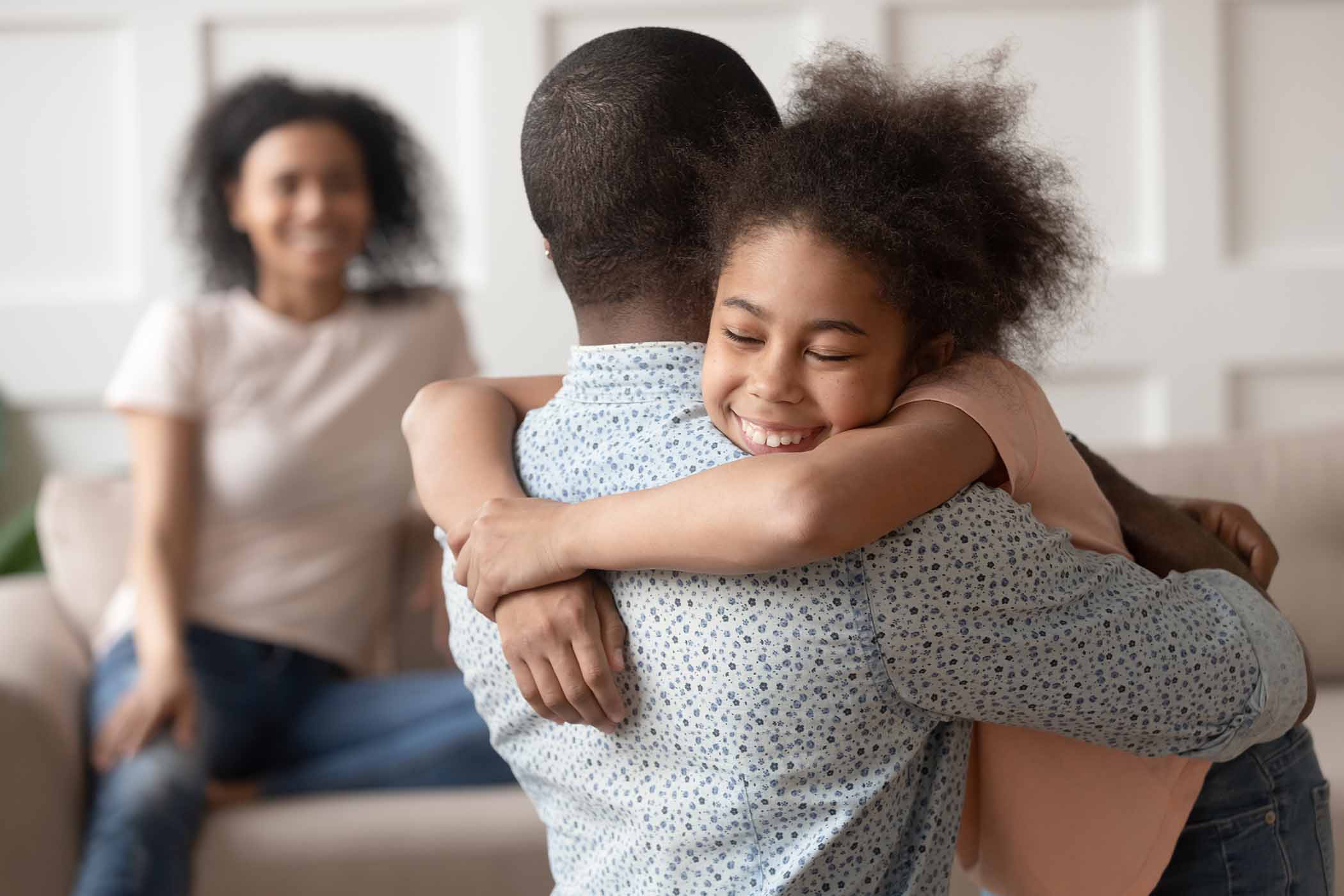 Happy cute little kid daughter embracing dad at home while the mother watches from the background with joy.